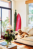 Light upholstered sofa with dog, cactus and surfboard in the corner of the room, in the foreground a coffee table with a bouquet of flowers