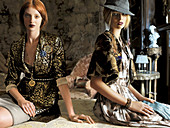 Zwei junge Frauen in Jacken, Kleid und Rock sitzen im wohnlichen Ambiente