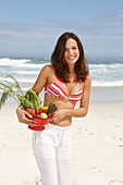 Junge brünette Frau mit Gemüseschale im Bikinoberteil und weißer Hose am Strand