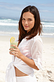 A young brunette woman on a beach with a smoothie wearing a white shirt and white trousers