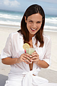 Junge brünette Frau mit Smoothie in weißem Hemd und weißer Hose am Strand