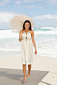 A brunette woman by the sea wearing a white summer dress and holding a parasol