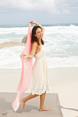 A brunette woman by the sea wearing a white summer dress and holding a pink shawl