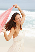 A brunette woman by the sea wearing a white summer dress and holding a pink shawl