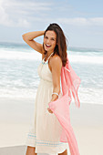 A brunette woman by the sea wearing a white summer dress and holding a pink shawl