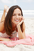 A brunette woman lying on a pink shawl by the sea wearing a white summer dress