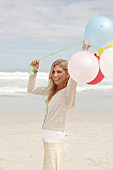 A blond woman on a beach wearing a light cardigan and shorts and holding balloons