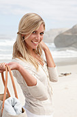 A blond woman on a beach wearing a light cardigan and holding a bag