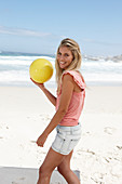 Junge Frau mit gelbem Ball im rosa Top und Jeansshorts am Strand