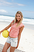 Junge Frau mit gelbem Ball im rosa Top und Jeansshorts am Strand