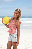 A young woman on a beach with a yellow ball wearing a pink top and denim shorts