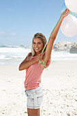 A young woman on a beach with balloons wearing a pink top and denim shorts