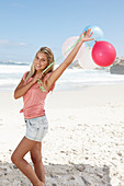 A young woman on a beach with balloons wearing a pink top and denim shorts