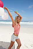 A young woman on a beach with balloons wearing a pink top and denim shorts