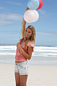 A young woman on a beach with balloons wearing a pink top and denim shorts