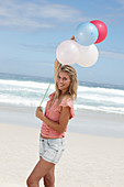 A young woman on a beach with balloons wearing a pink top and denim shorts