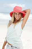 A blonde woman on the beach wearing a light t-shirt and a red hat