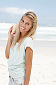 A blonde woman on the beach wearing a light t-shirt and holding a shell