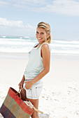 A blonde woman on the beach wearing a light t-shirt and denim shorts holding a wicker bag