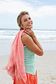 A blonde woman on a beach with a scarf wearing a turquoise top and a salmon-pink skirt