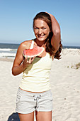 A brunette woman on a beach with a slice of watermelon wearing a light top and shorts