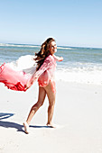 A brunette woman with a beach towel wearing a pink blouse and a bikini by the sea