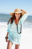 A brunette woman wearing a hat, a necklace and a light-blue beach dress by the sea