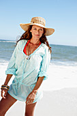 A brunette woman wearing a hat and a light-blue beach dress by the sea