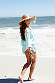 A brunette woman wearing a hat and a light-blue beach dress by the sea