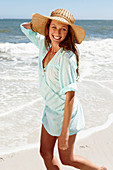 A brunette woman wearing a hat and a light-blue beach dress by the sea