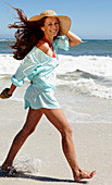 A brunette woman wearing a hat and a light-blue beach dress by the sea