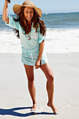 A brunette woman wearing a hat and a light-blue beach dress by the sea