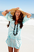 A brunette woman wearing a hat, a necklace and a light-blue beach dress by the sea