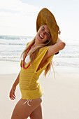 A blonde woman on a beach wearing a hat, a yellow knitted top and a bikini