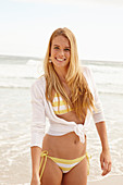 A blonde woman on a beach wearing a white and yellow bikini and a cardigan