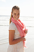 A blonde woman by the sea wearing a salmon-pink scarf and a white dress