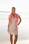 A blonde woman by the sea wearing a salmon-pink scarf and a white dress