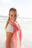 A blonde woman by the sea wearing a salmon-pink scarf and a white dress