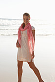 A blonde woman by the sea wearing a salmon-pink scarf and a white dress