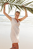 A blonde woman by the sea holding a palm frond wearing a white dress