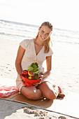 A blonde woman by the sea with a bowl of fruit and vegetables wearing a white dress