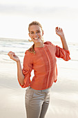 A young blonde woman wearing an orange blouse and light trousers by the sea