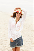 A brunette woman wearing a hat, a white blouse and denim shorts