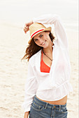 A brunette woman wearing a hat, a red bikini top, a white blouse and denim shorts