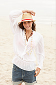 A brunette woman wearing a hat, a white blouse and denim shorts