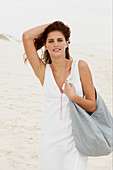 A brunette woman by the sea with a bag wearing a white summer dress