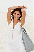 A brunette woman by the sea with a bag wearing a white summer dress