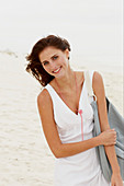 A brunette woman by the sea with a bag wearing a white summer dress