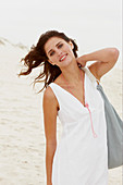 A brunette woman by the sea with a bag wearing a white summer dress