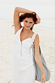 A brunette woman by the sea with a bag wearing a white summer dress